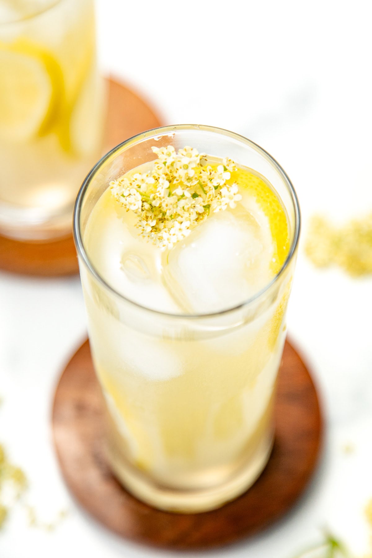 A glass of iced lemonade with lemon slices and small white flowers, placed on a wooden coaster.