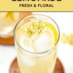 A glass of elderflower lemonade with ice and lemon, topped with elderflowers, text promoting the recipe.