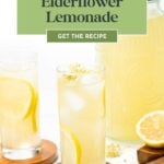 Two glasses of elderflower lemonade with lemon slices, a pitcher, and flowers on a white surface.