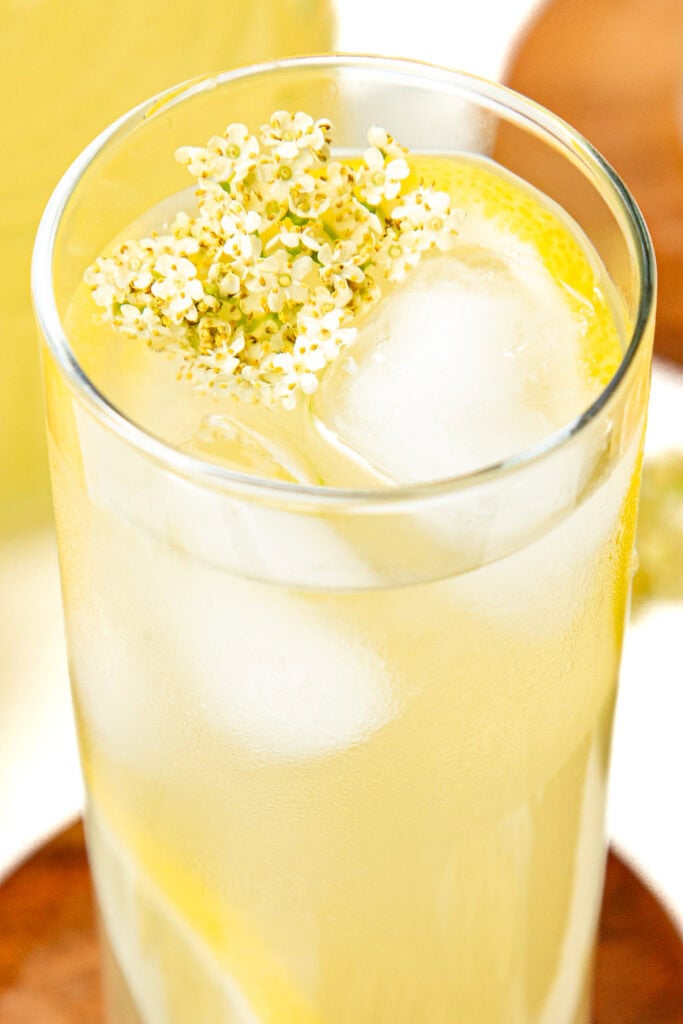 A glass of lemonade with ice, lemon slices, and a small cluster of white elderflower blossoms on top.