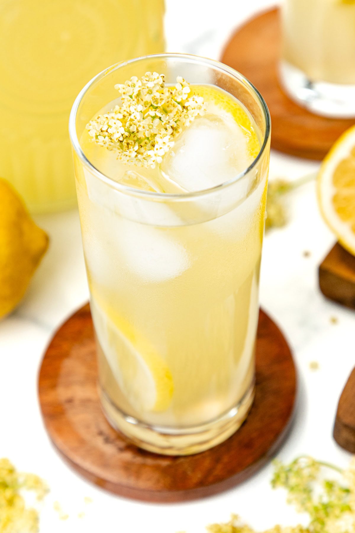 A tall glass of iced lemonade with lemon slices and white flowers, on a wooden coaster.