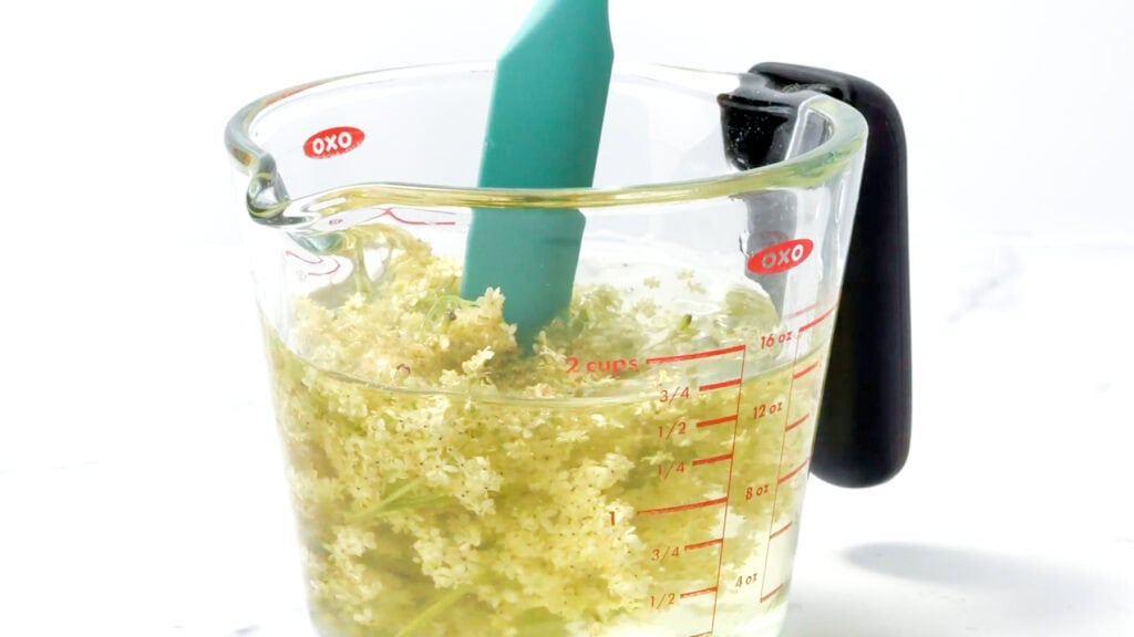 Elderflowers and lemonade combine in a glass measuring cup with a teal spatula, resting on a white surface.