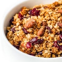 A bowl of granola with oats, almonds, and dried cranberries on a white background.