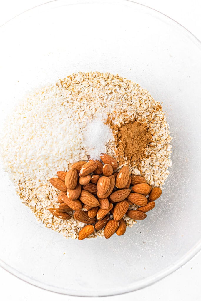 A glass bowl with oats, whole almonds, shredded coconut, cinnamon, and sugar on a white background.