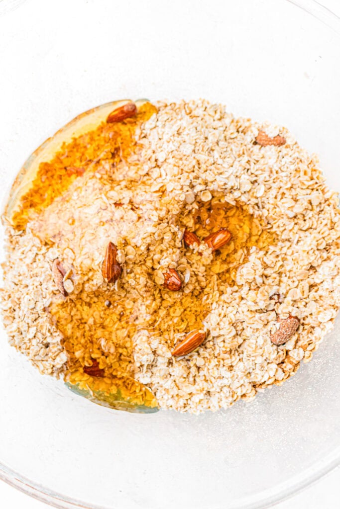 Oats, sliced almonds, and honey are mixed together in a clear glass bowl.