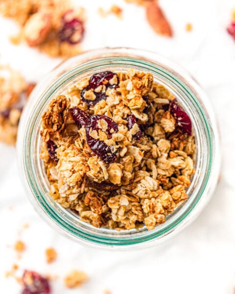 A glass jar filled with granola clusters, oats, and dried cranberries on a white surface.