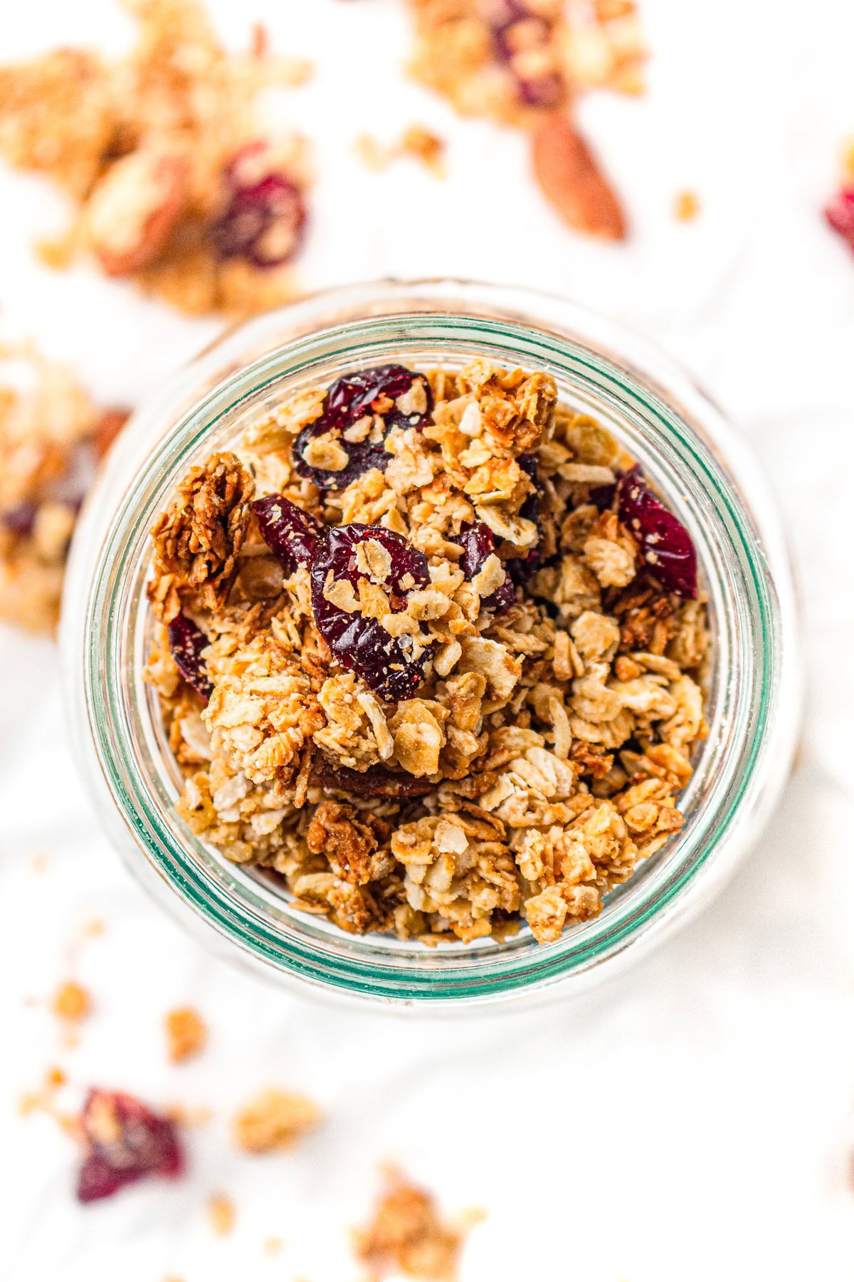A glass jar filled with granola clusters, oats, and dried cranberries on a white surface.