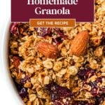 Close-up of homemade granola with almonds and dried cranberries in a white bowl, recipe text overlayed.