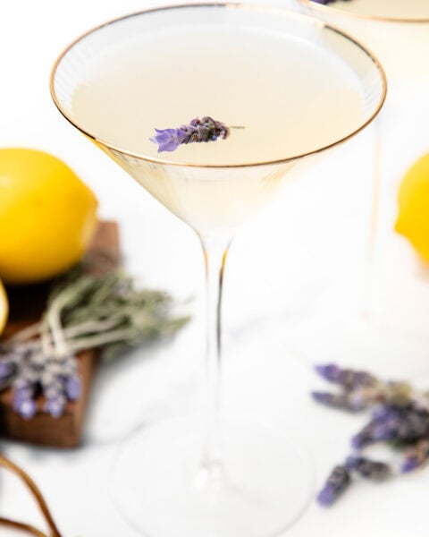 A martini glass with a pale cocktail garnished with lavender, lemons, and more lavender in the background.