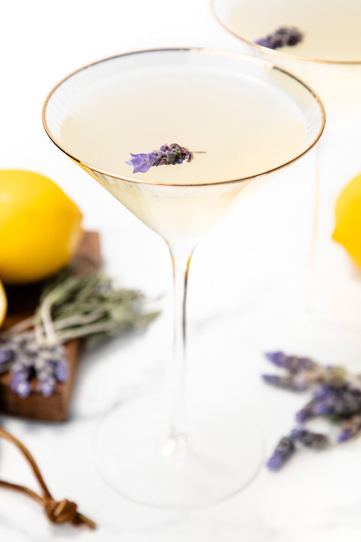 A martini glass with a pale cocktail garnished with lavender, lemons, and more lavender in the background.