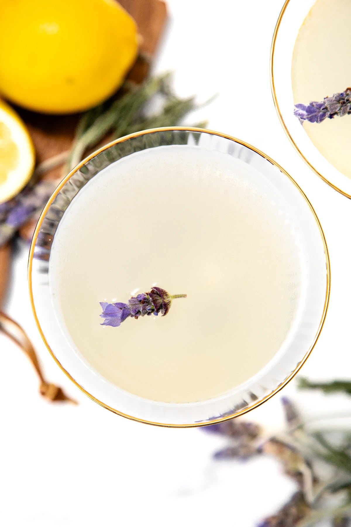A cocktail in a glass garnished with a sprig of lavender, with lemons and herbs in the background.