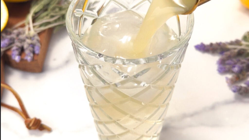 Pale yellow liquid being poured into a crystal glass filled with ice, on a white surface.