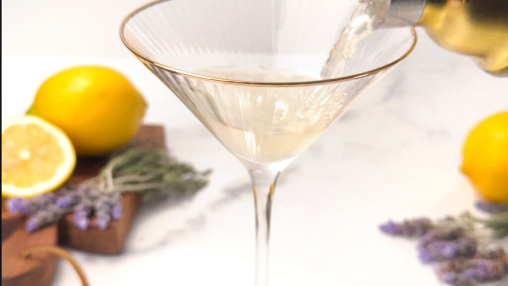 Champagne being poured into a martini glass with lemons and lavender on a marble table in the background.