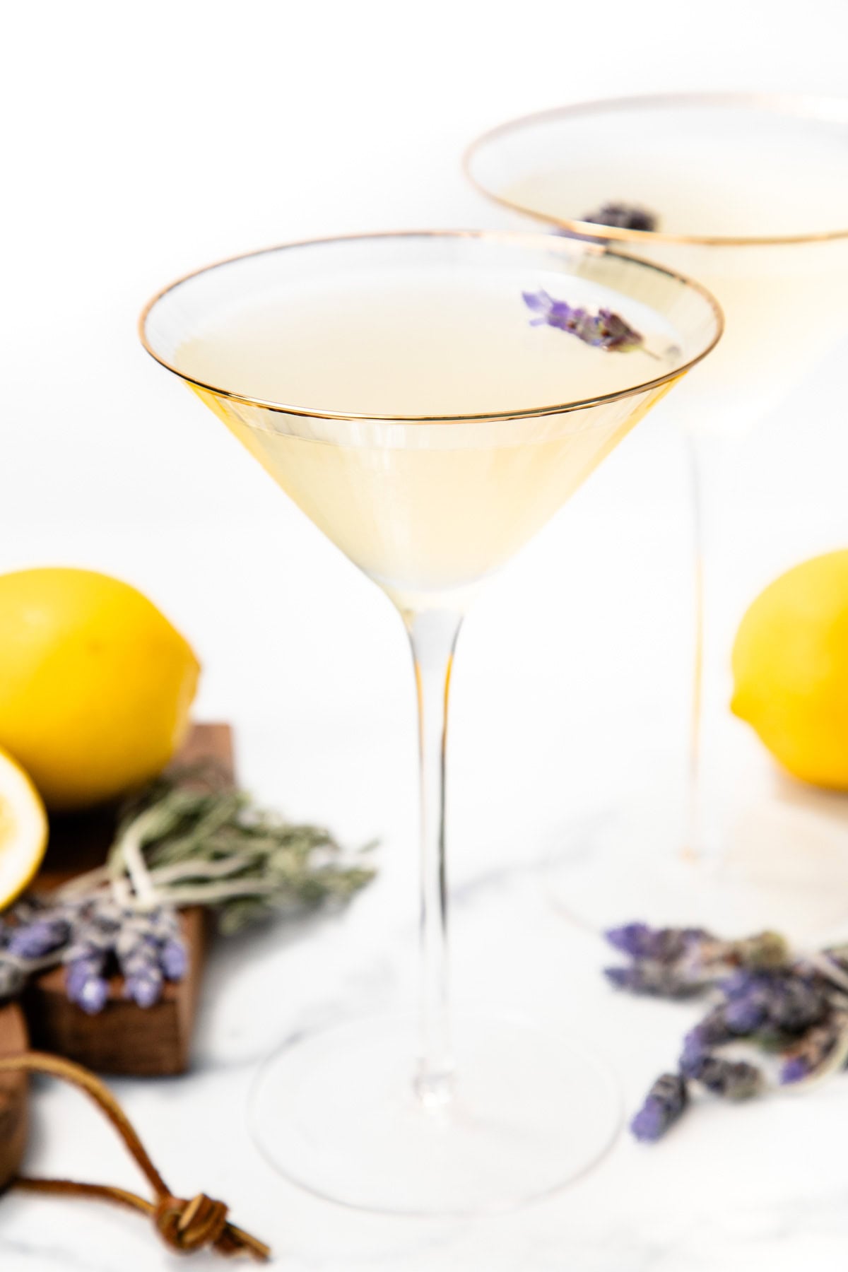 A pale yellow cocktail in a martini glass, garnished with lavender, with lemons and lavender sprigs nearby.