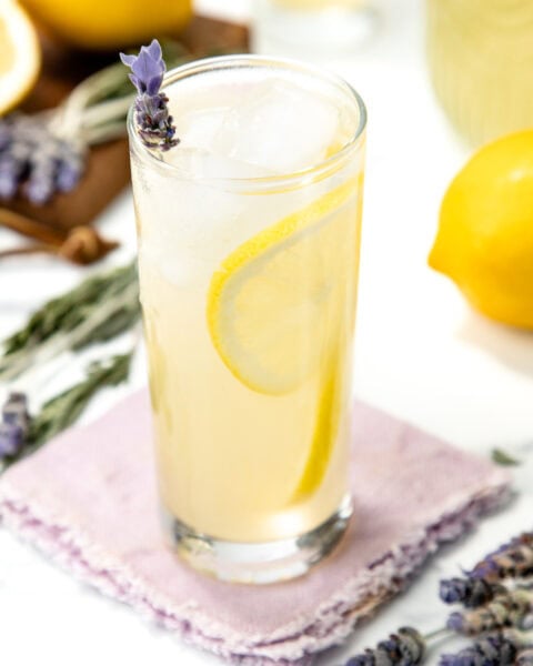 Tall glass of lemonade with lemon slice, ice, and lavender sprig on a purple napkin, lemons nearby.