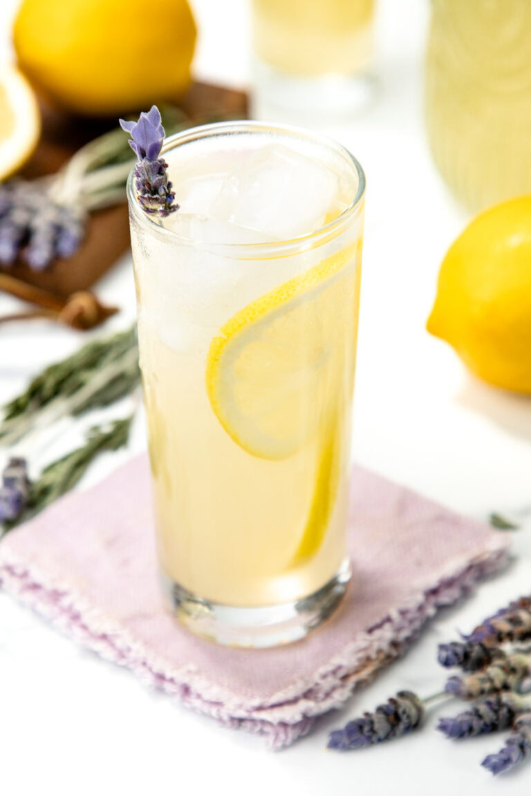Tall glass of lemonade with lemon slice, ice, and lavender sprig on a purple napkin, lemons nearby.