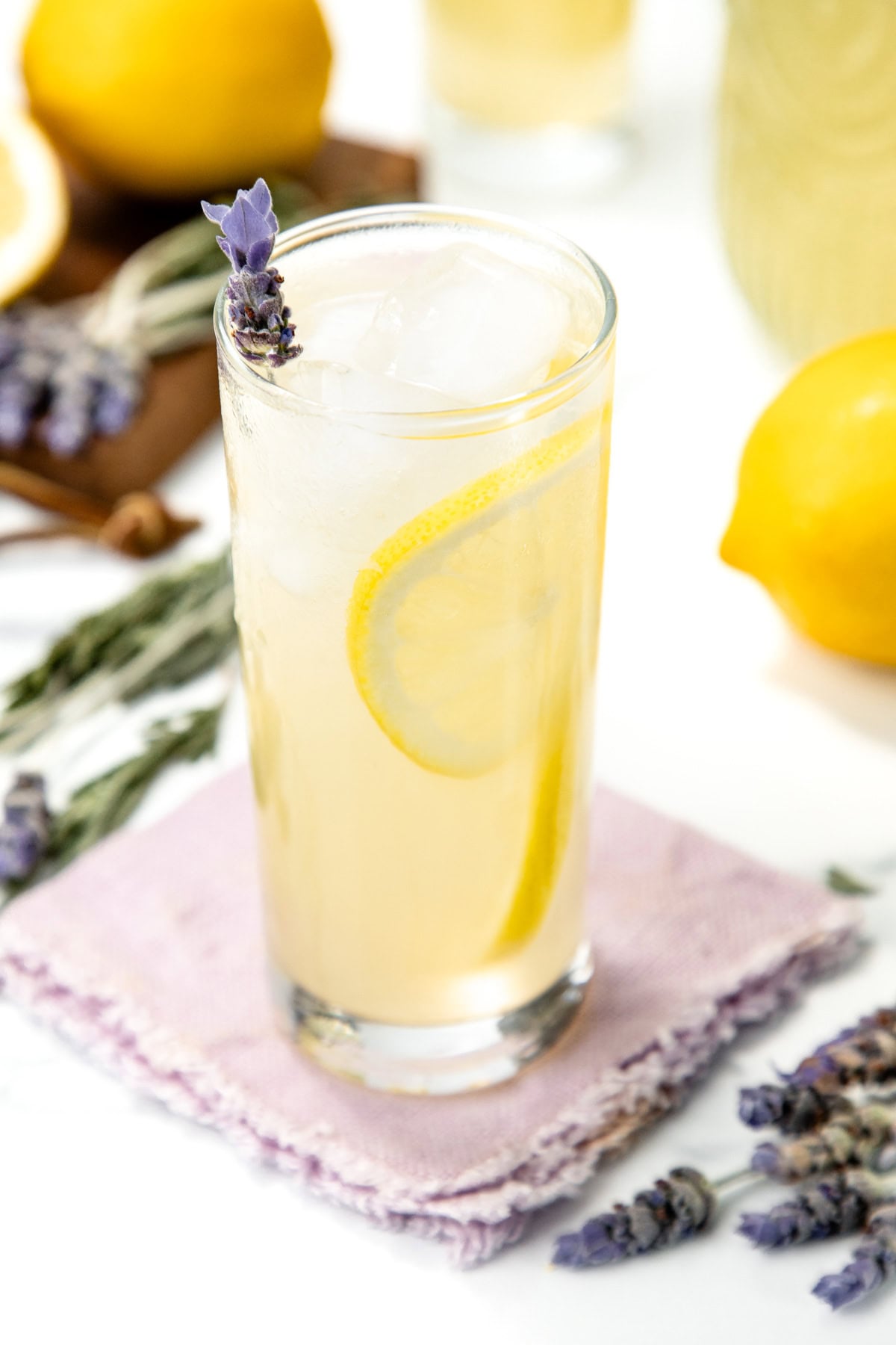 Tall glass of lemonade with lemon slice, ice, and lavender sprig on a purple napkin, lemons nearby.