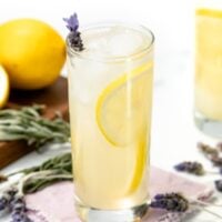 Iced lemonade with lemon slice and lavender sprig, with lemons and lavender on a table in the background.
