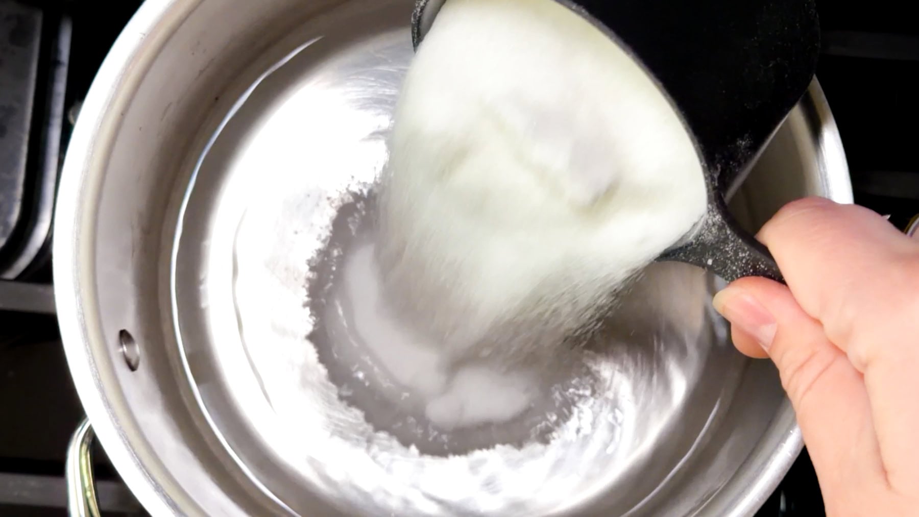 Hand pouring white sugar from a measuring cup into a metal pot on a stove, viewed from above.