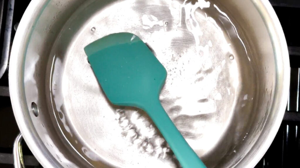 A teal silicone spatula rests in a pot of clear liquid on a stovetop.