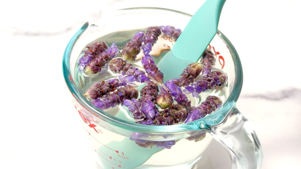 Clear measuring cup with water, purple dried flowers, and a teal spatula stirring inside.