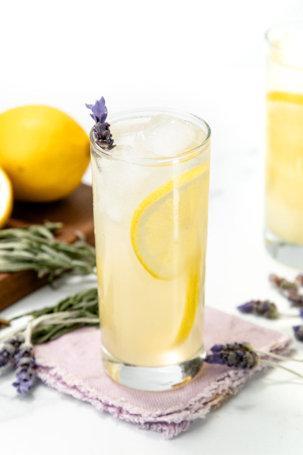 A tall glass of lemonade with ice, lemon slices, and a sprig of lavender, next to fresh lemons.