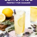 Glass of lavender lemonade with lemon slices, lavender sprigs, and lemons on a white table.