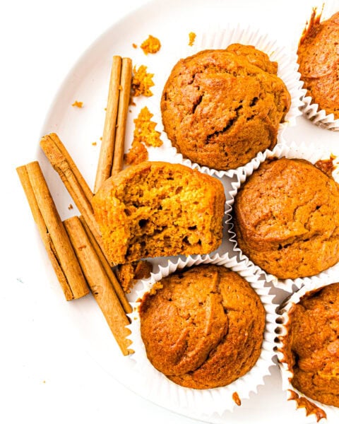 Pumpkin muffins on a white plate with cinnamon sticks and crumbs, one muffin is cut in half.