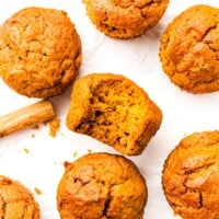 Six pumpkin muffins on white paper, one with a bite taken out, and a cinnamon stick beside them.