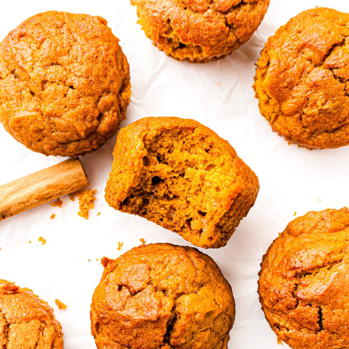 Six pumpkin muffins on white paper, one with a bite taken out, and a cinnamon stick beside them.