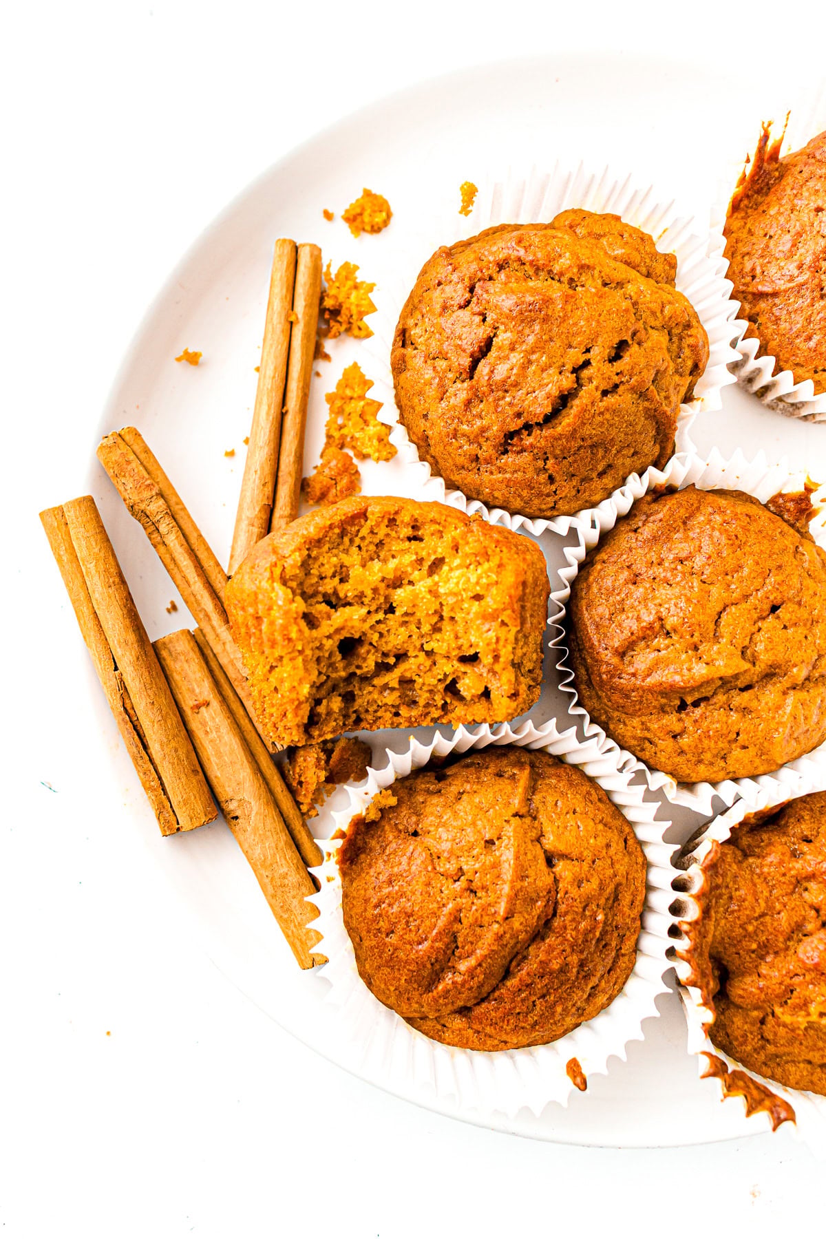Pumpkin muffins on a white plate with cinnamon sticks and crumbs, one muffin is cut in half.