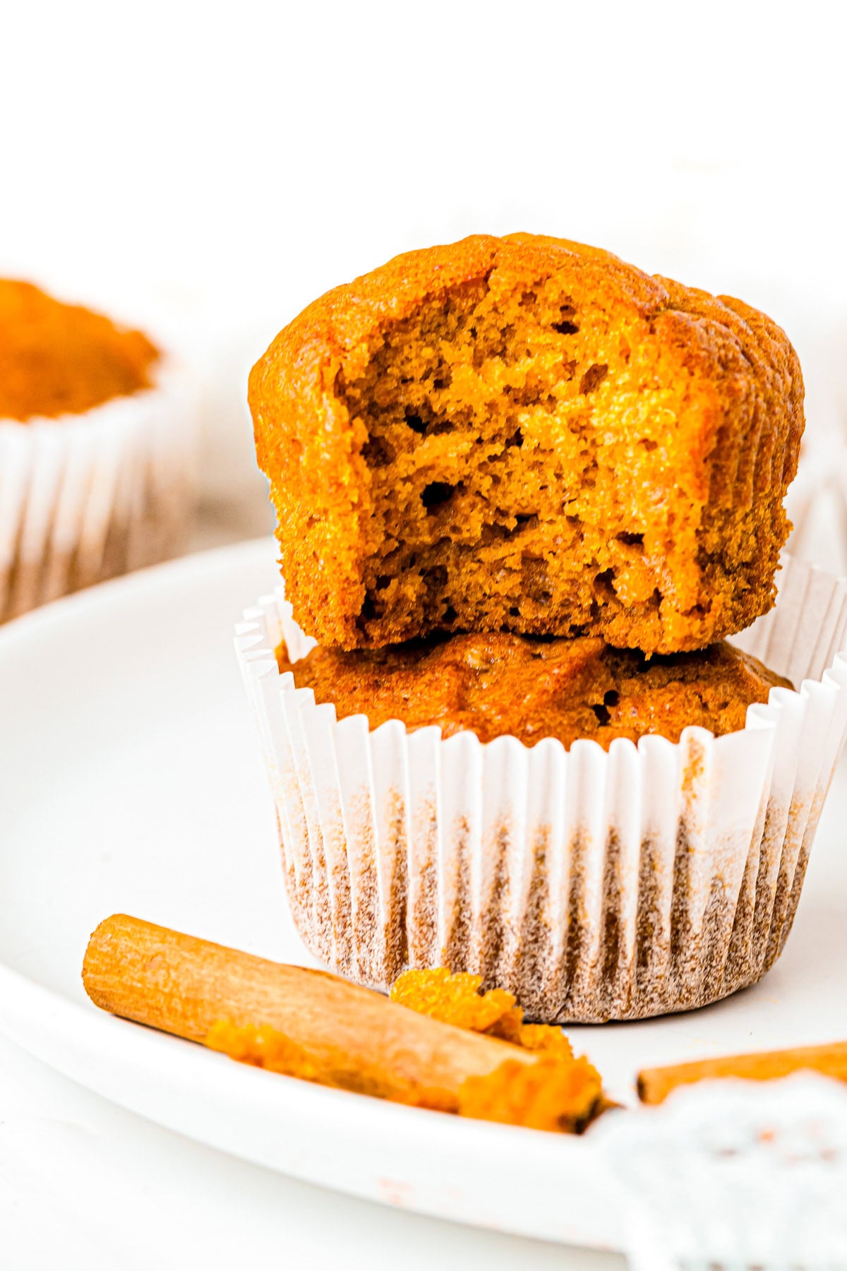 Two pumpkin muffins in white liners, one is halved and stacked on top, with a cinnamon stick nearby.