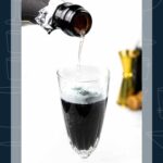 A bottle pours champagne into a tall glass filled with black liquid, with bar tools in the background.