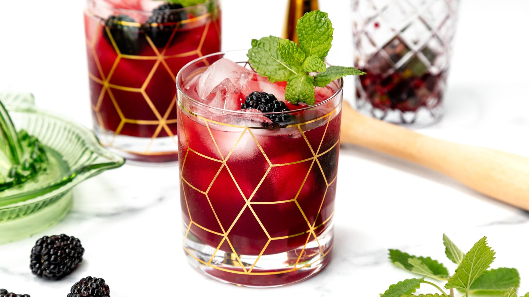 A glass of blackberry cocktail with ice, mint garnish, and gold geometric pattern, on a white surface.