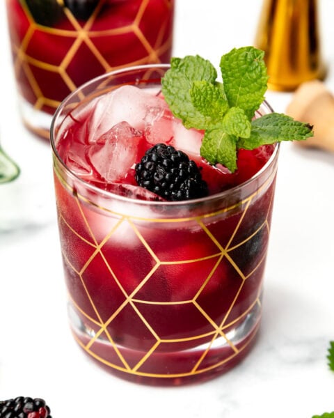 A glass of berry cocktail with ice, blackberry, and mint garnish, in a geometric gold-patterned glass.