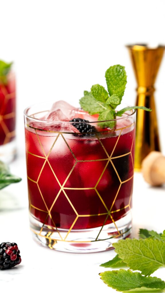 A glass of blackberry cocktail with ice, mint garnish, and gold geometric pattern on the glass.
