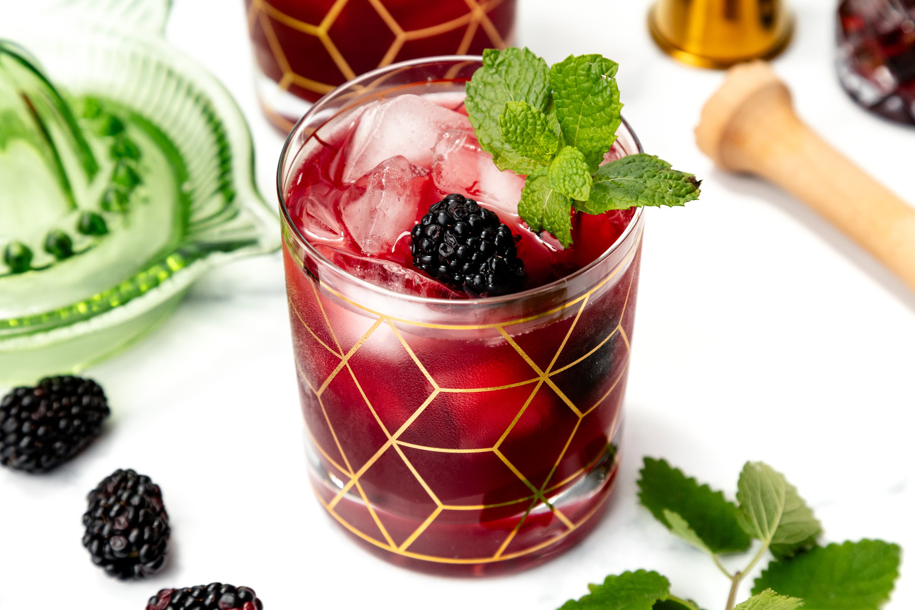 A glass of berry cocktail with ice, garnished with mint and blackberry, sits on a white surface.