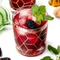 A cocktail with ice, blackberry, and mint garnish in a glass with a gold geometric pattern.