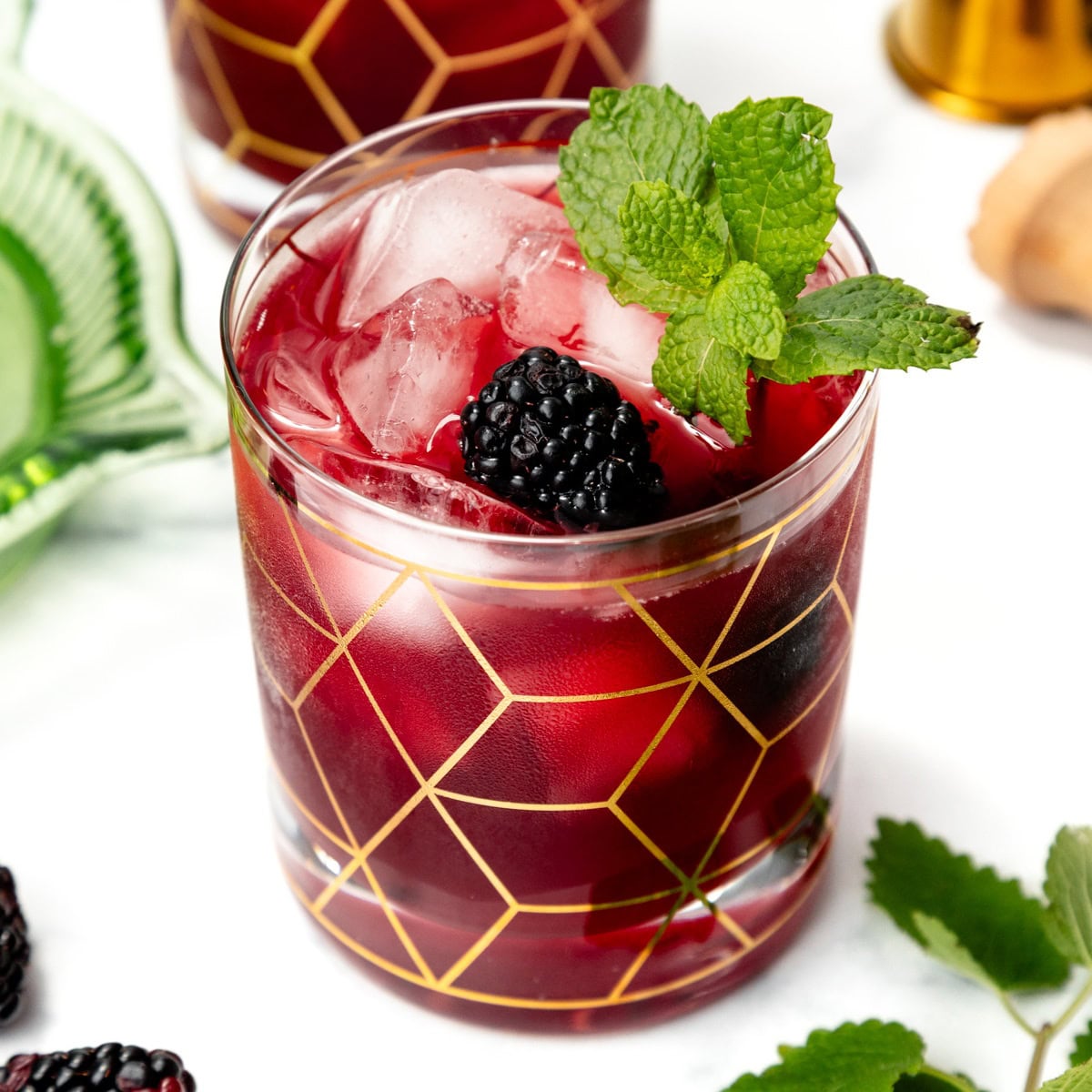 A cocktail with ice, blackberry, and mint garnish in a glass with a gold geometric pattern.