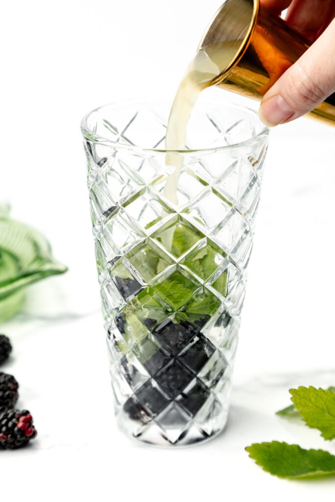 A hand pours liquid into a glass with mint leaves and berries at the bottom.