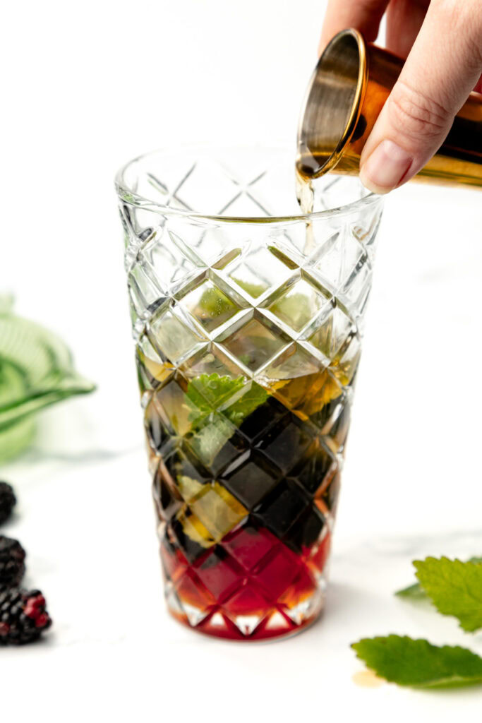 A hand pours liquid into a tall glass with ice, mint leaves, and red and dark layers, on a white surface.