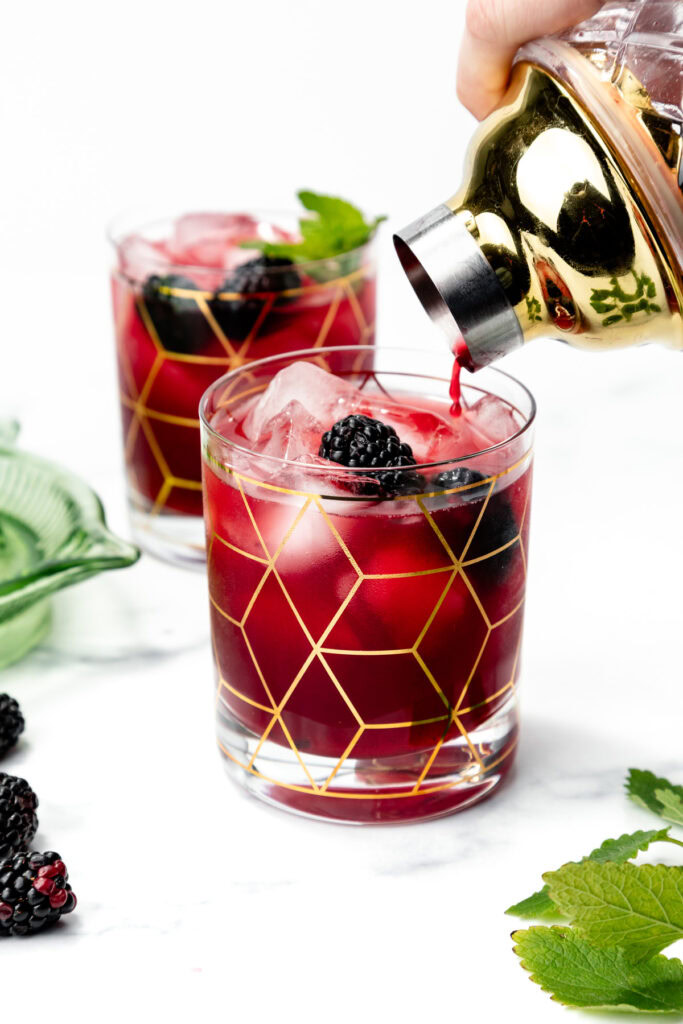 A hand pours a red blackberry cocktail into a glass with gold geometric patterns and ice.