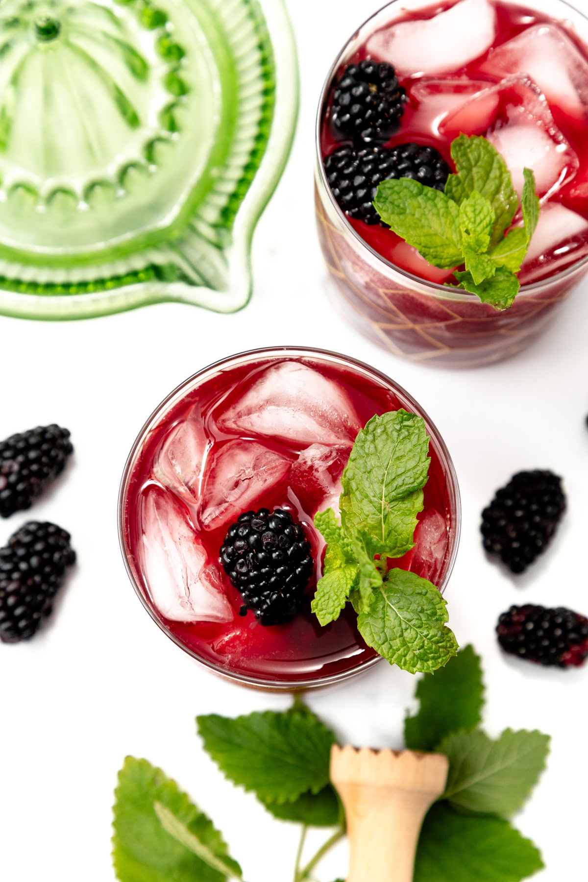Two glasses of blackberry drink with ice, mint, and blackberries, surrounded by fresh blackberries and leaves.