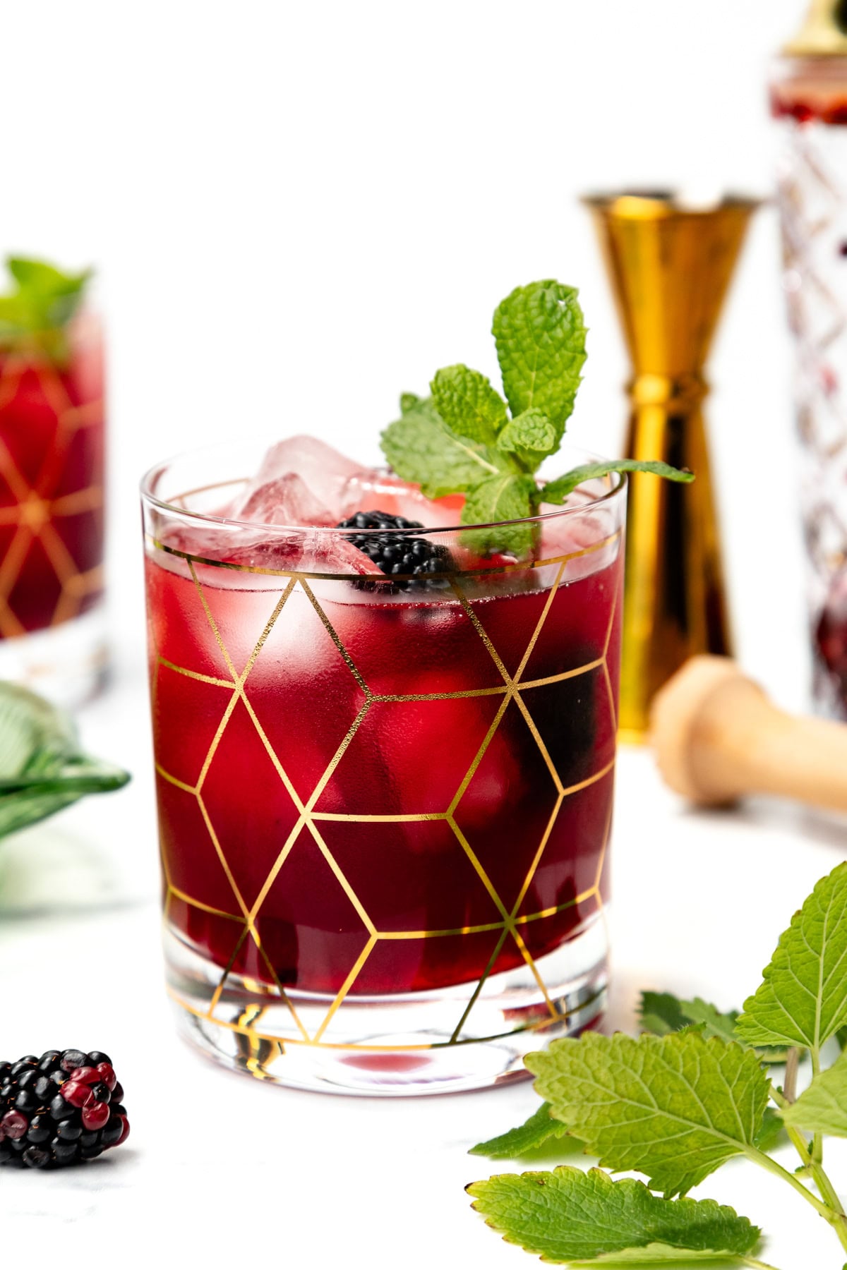 A purple cocktail with ice, blackberry, and mint in a geometric glass, surrounded by bar tools.