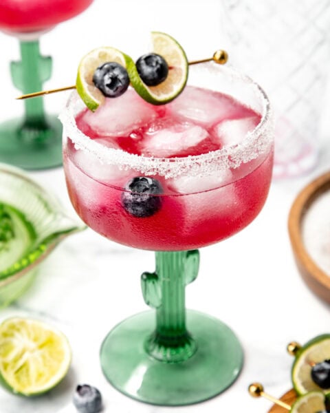 A pink cocktail with ice, blueberries, and lime garnish in a cactus glass, with salt on the rim.