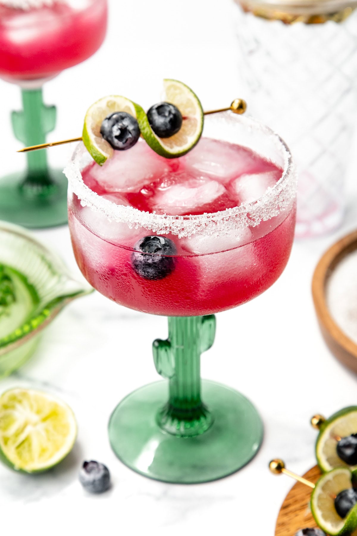 A pink cocktail with ice, blueberries, and lime garnish in a cactus glass, with salt on the rim.