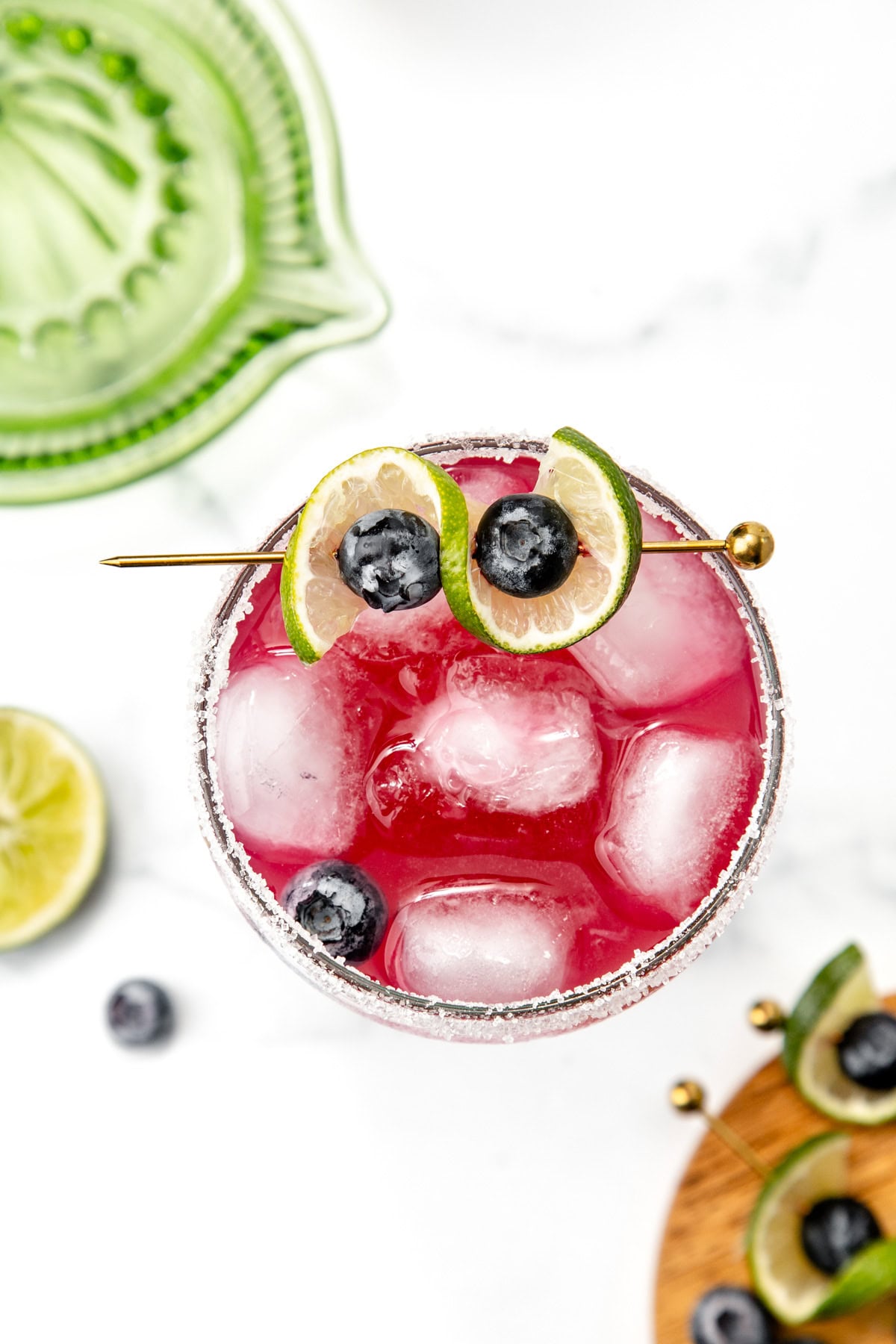 A pink cocktail with ice, garnished with lime slices and blueberries on a skewer, seen from above.