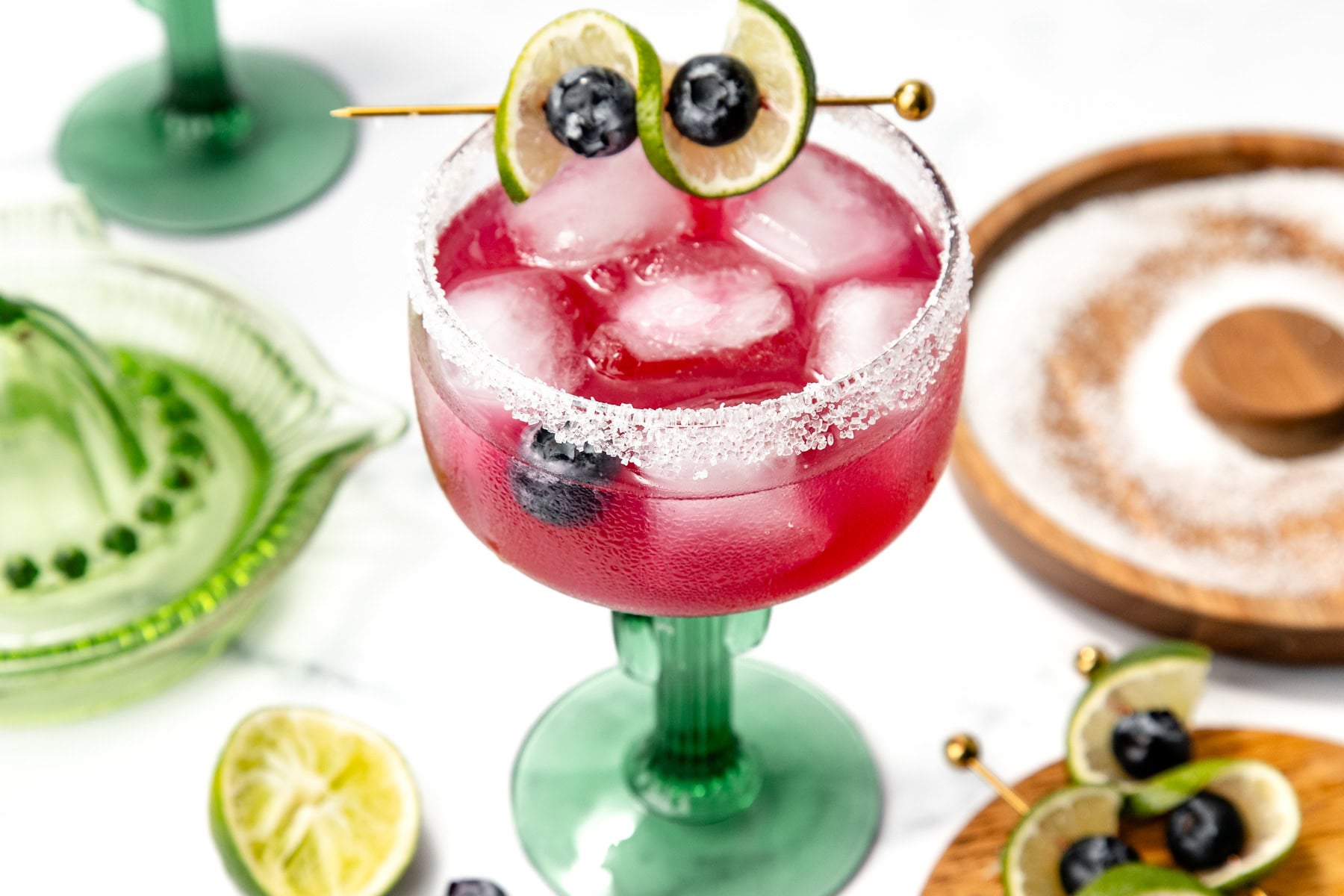A pink margarita with ice, lime, and blueberries in a green glass with a salted rim on a white background.