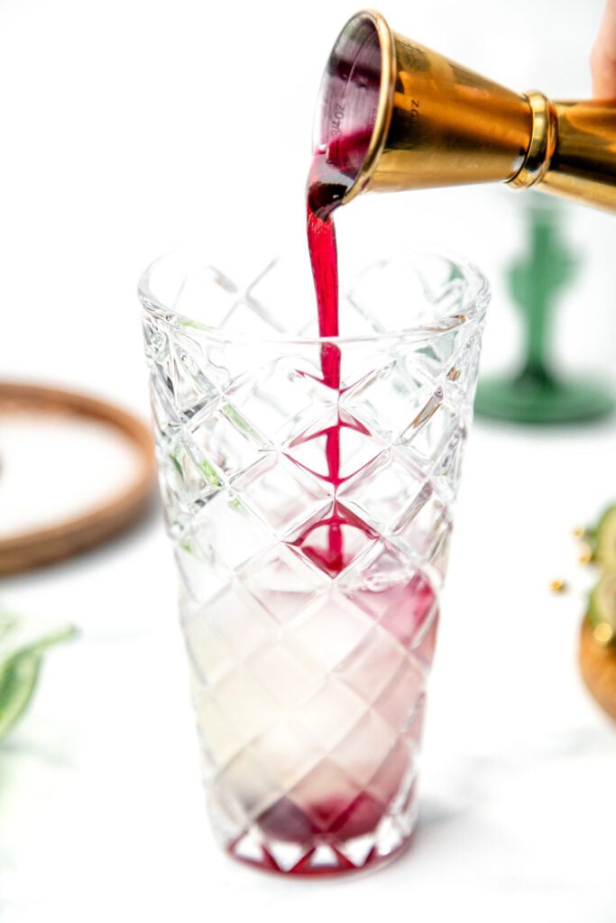 Red liquid being poured from a jigger into a tall, patterned glass with white liquid inside.