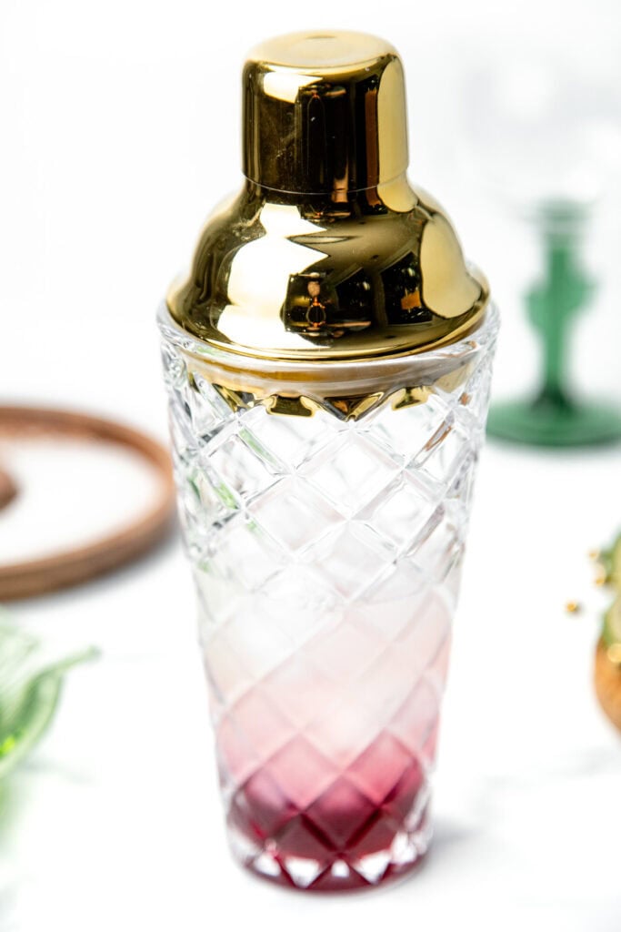 Crystal cocktail shaker with a gold lid, containing a small amount of red liquid, on a white background.